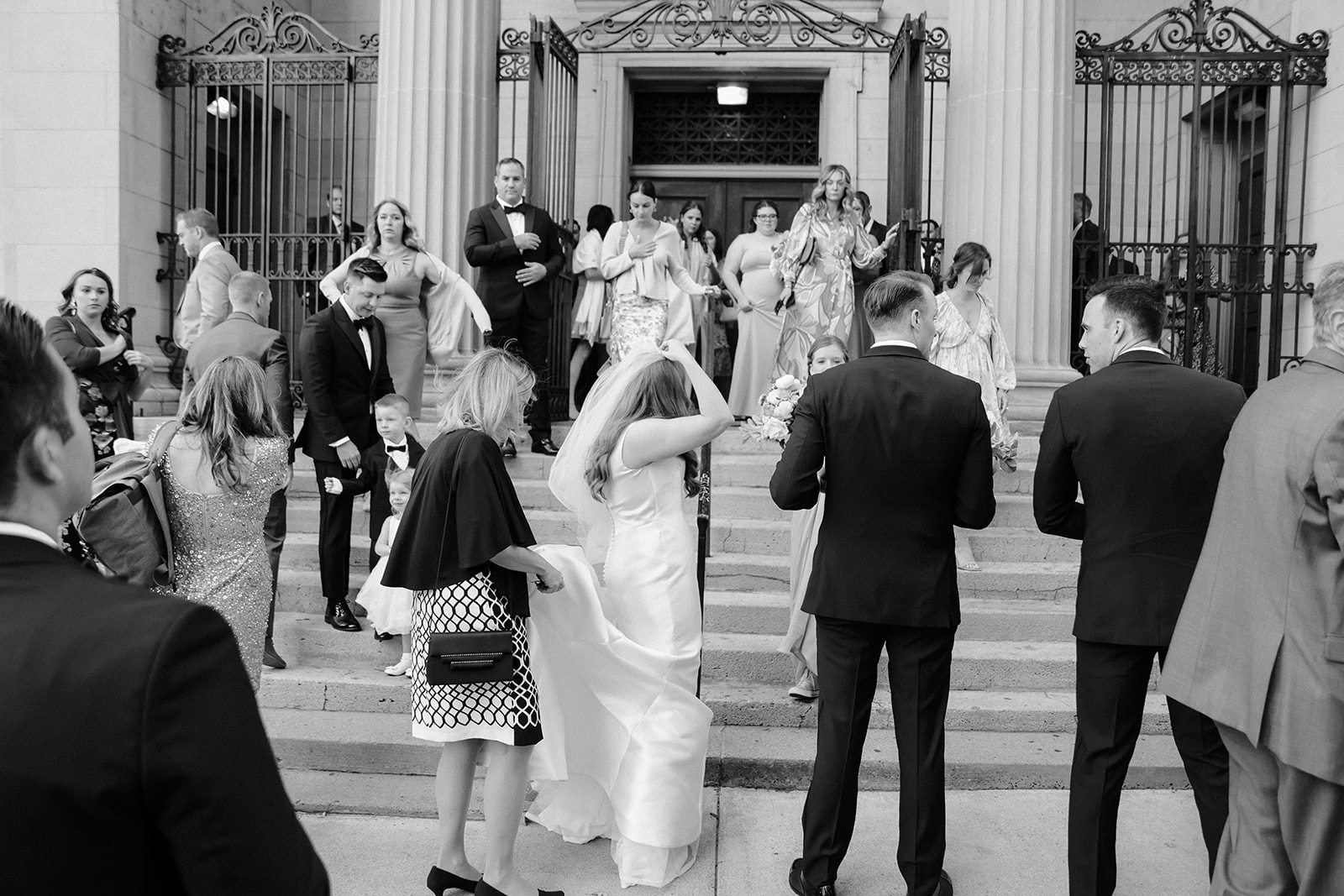 Candid capture of a busy wedding party outside the steps of their venue as the bride adjusts her veil.